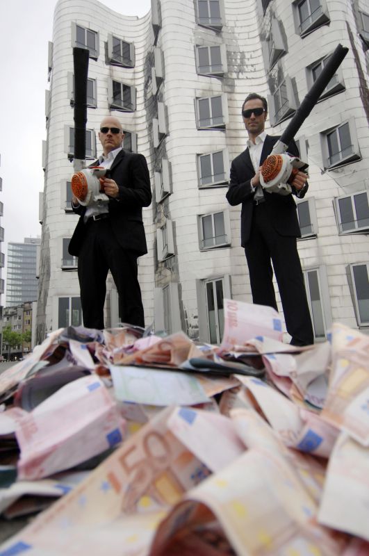 Männer im schwarzen Anzug mit Laubbläsern vor einem Haufen Geldscheinen