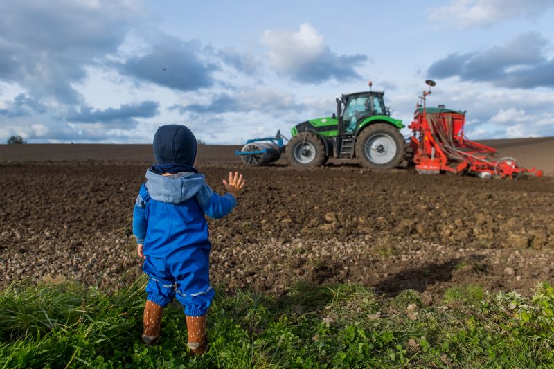 Kind steht am Rande eines Ackers und winkt Traktor zu