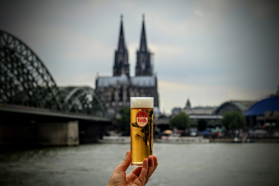 Eine Hand hält ein Glas Kölsch, im Hintergrund der Kölner Dom