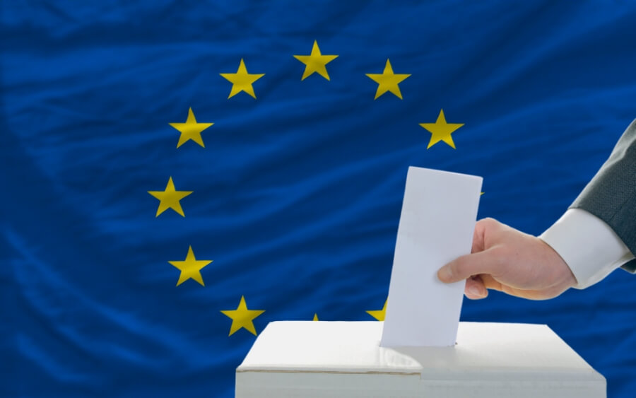 Eine Person steckt einen Zettel in eine Wahlurne, im Hintergrund die Flagge der EU