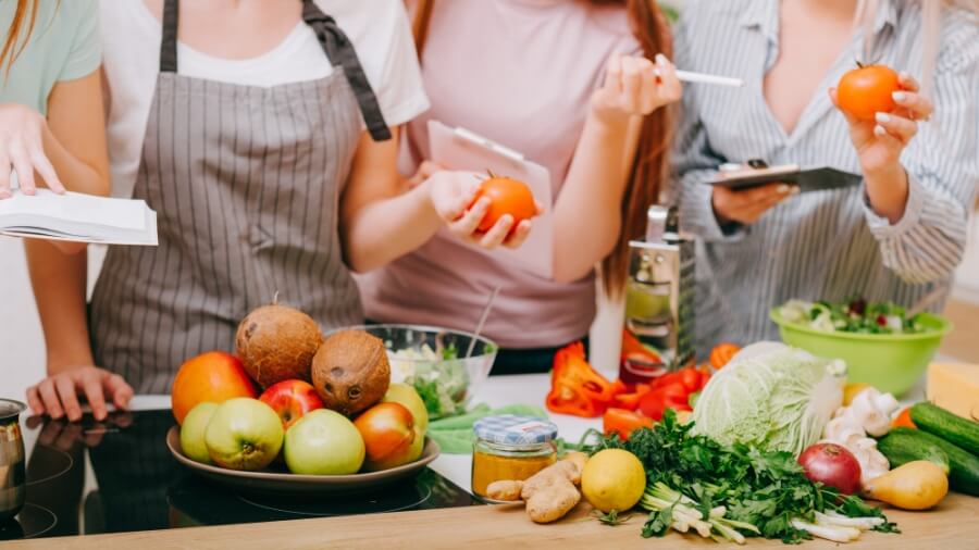 Personen in einem Kochkurs stehen vor einer Arbeitsfläche mit Obst und Gemüse