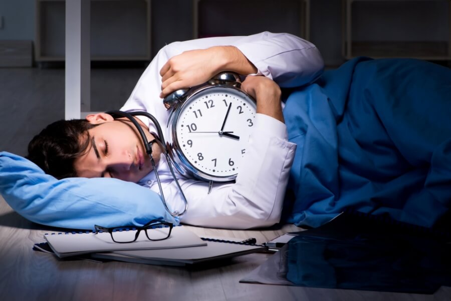 Junger Arzt schläft mit Uhr in der Hand auf dem Boden. Symbolbild Überstunden.
